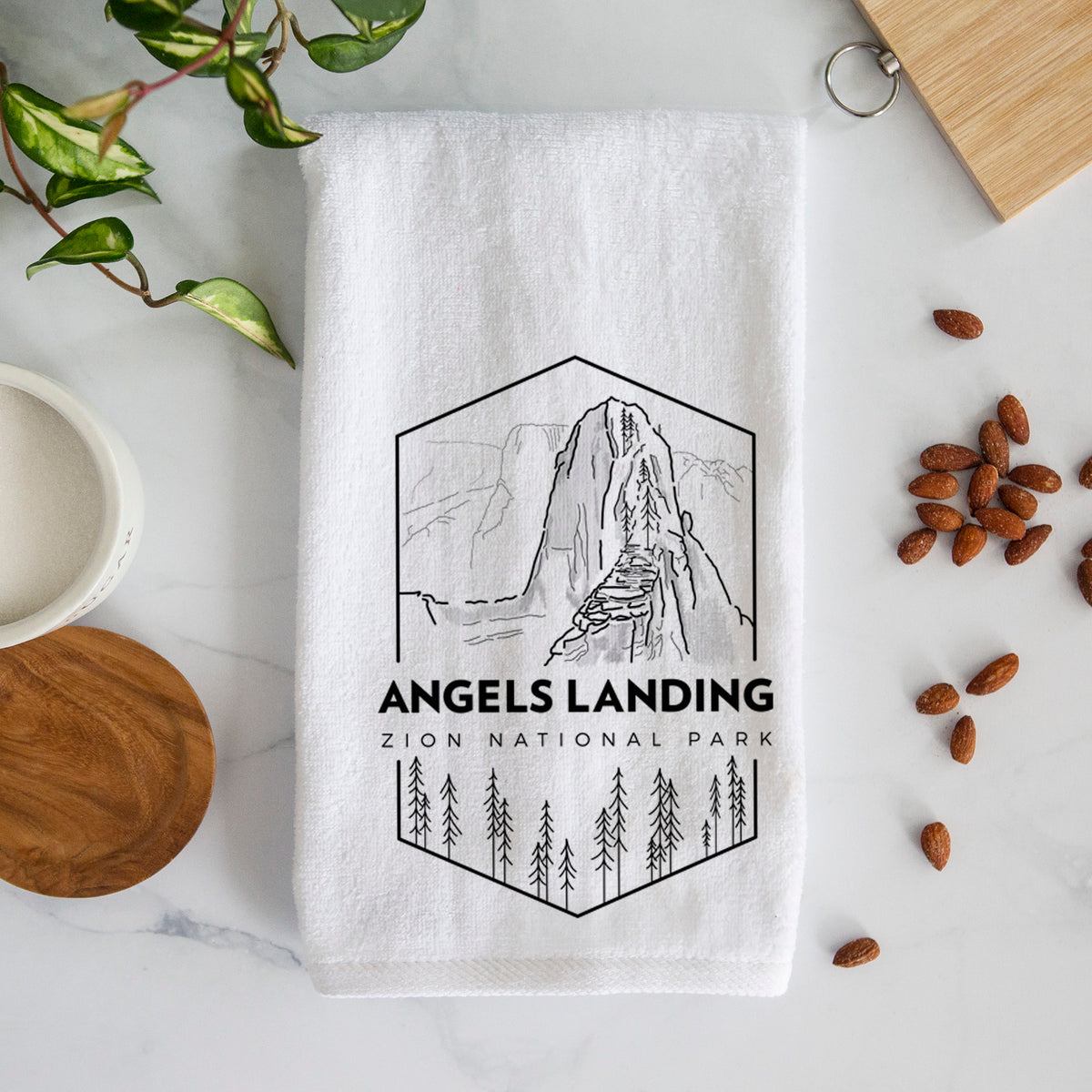 White hand towel printed with a black line art badge of Angels Landing above pine trees, with the words Angels Landing, Zion National Park.
