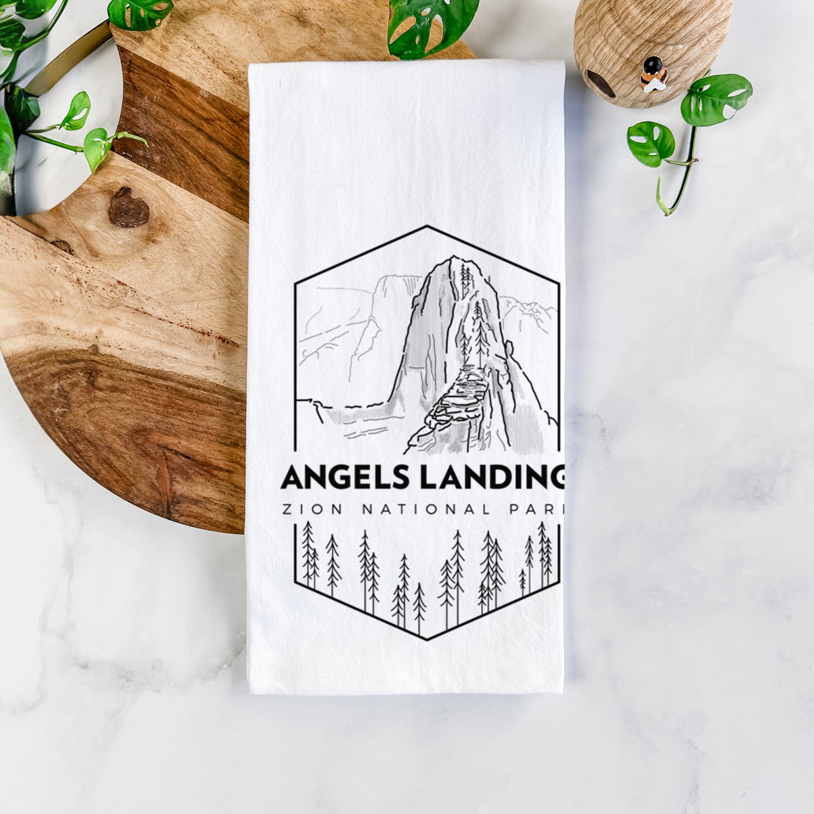 White cotton tea towel printed with a black line art badge of Angels Landing above pine trees, with the words Angels Landing, Zion National Park, on a marble counter beside a round wooden cutting board and greenery.
