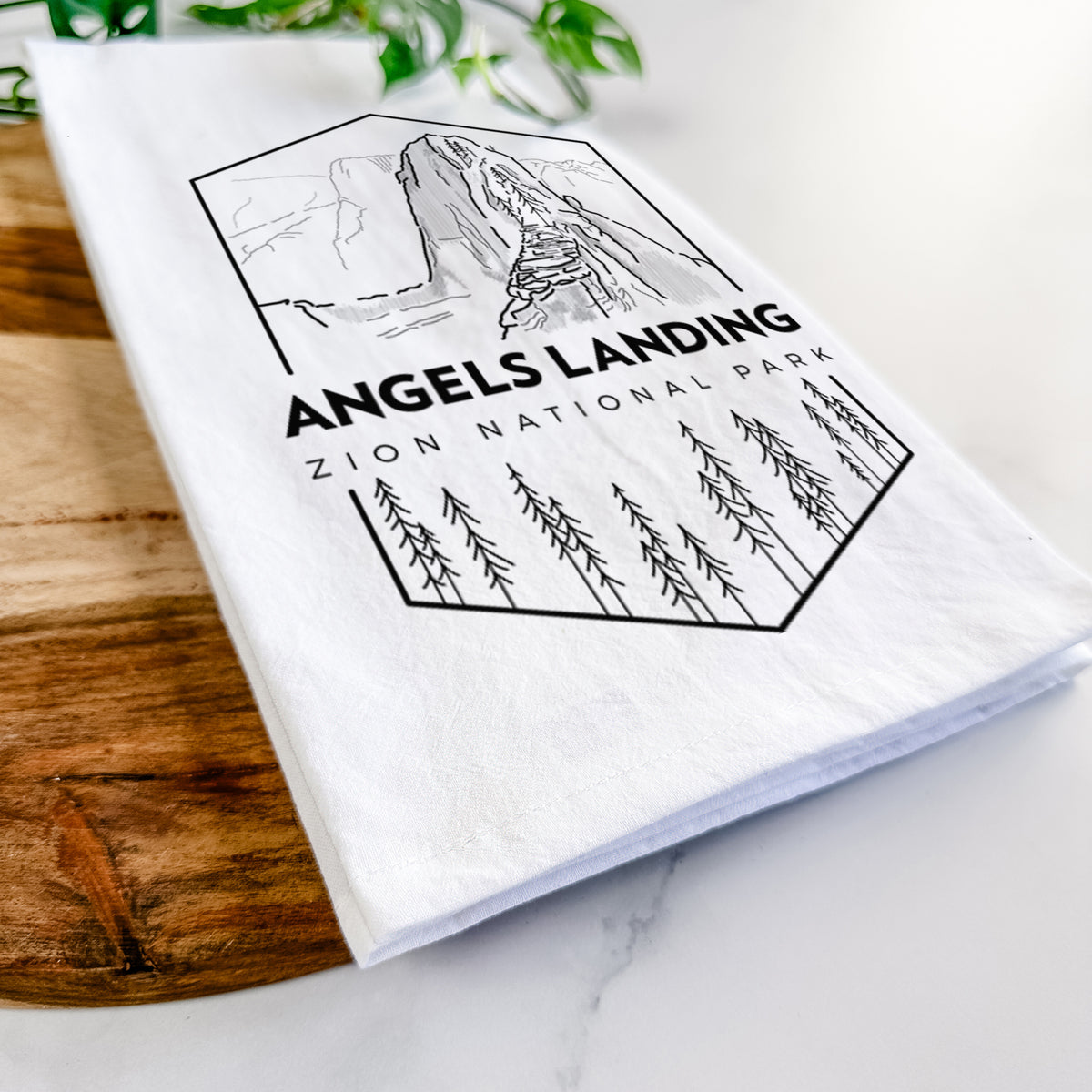 Close-up of a folded white tea towel on a wooden board, showing a black line art badge of Angels Landing above pine trees, with the words Angels Landing, Zion National Park.