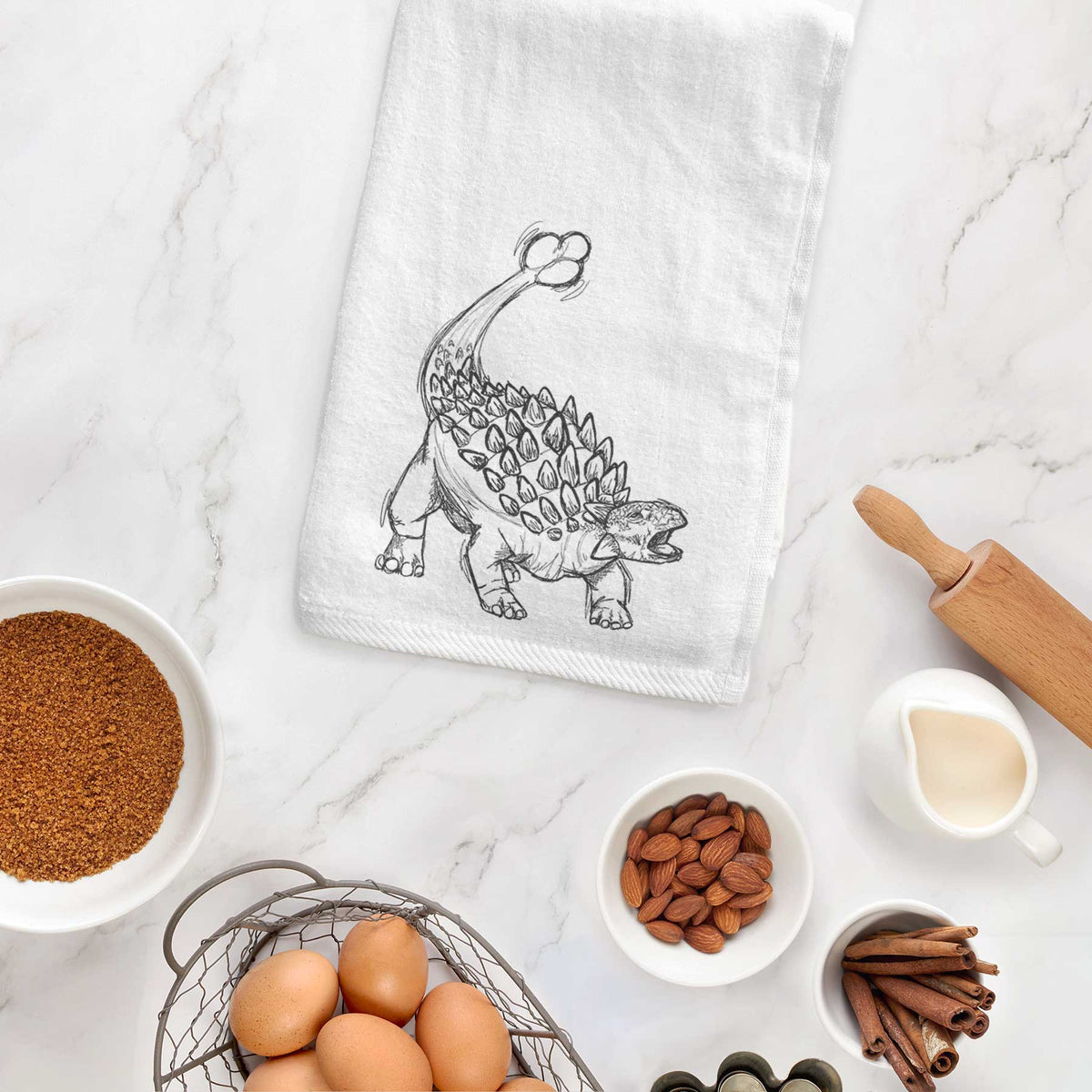 White hand towel printed with a black ink illustration of an Ankylosaurus with its club tail raised, hanging on a cabinet door.