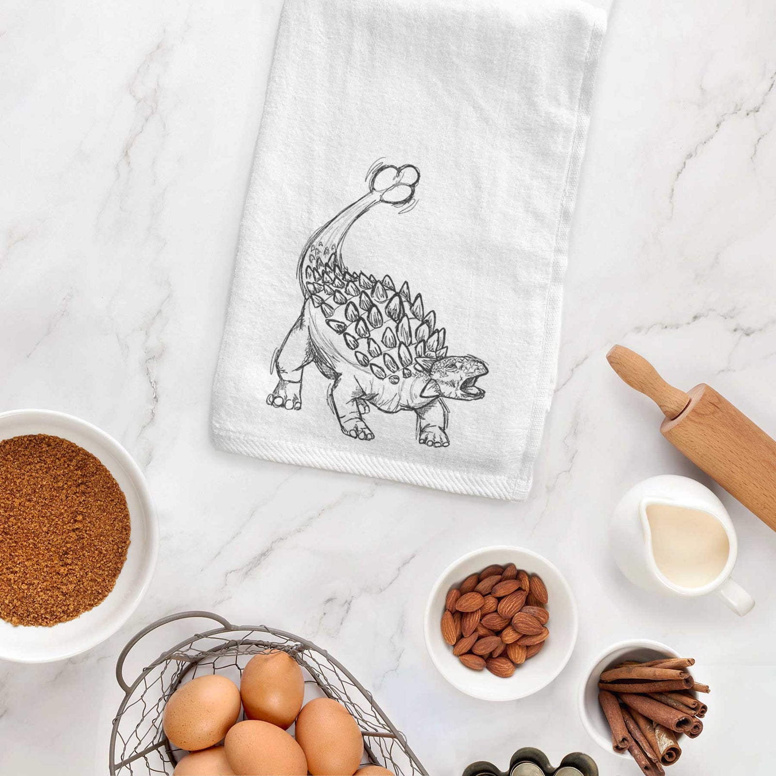 White hand towel printed with a black ink illustration of an Ankylosaurus with its club tail raised, hanging on a cabinet door.