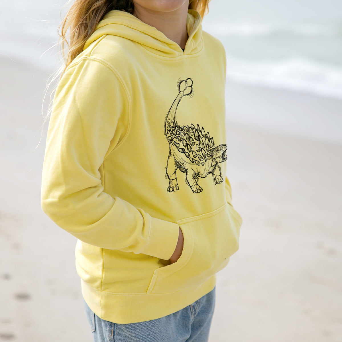 Girl wearing a youth pigment dyed hoodie printed with a black ink illustration of an Ankylosaurus with its club tail raised.