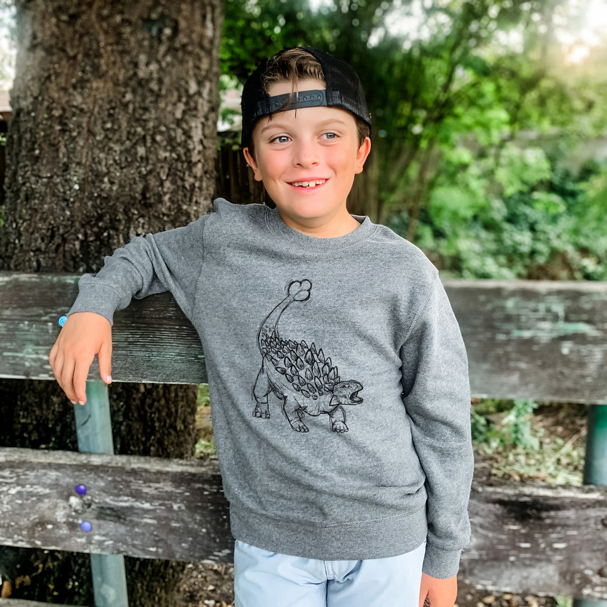 Boy wearing a youth crewneck sweatshirt printed with a black ink illustration of an Ankylosaurus with its club tail raised, outdoors.