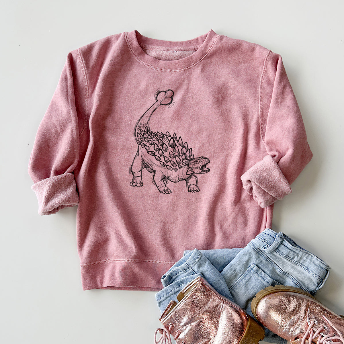 Youth crewneck sweatshirt printed with a black ink illustration of an Ankylosaurus with its club tail raised, styled flat with jeans and boots.