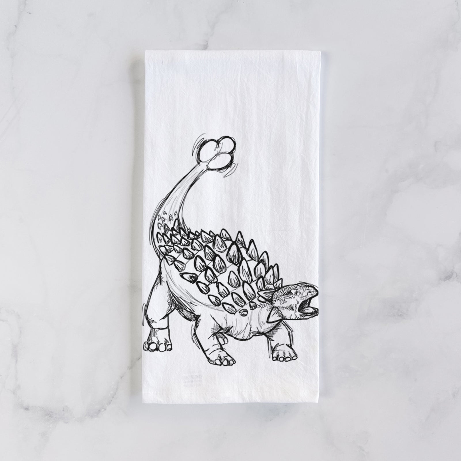 White cotton tea towel printed with a black ink illustration of an Ankylosaurus with its club tail raised, on a marble counter beside a round wooden cutting board and greenery.
