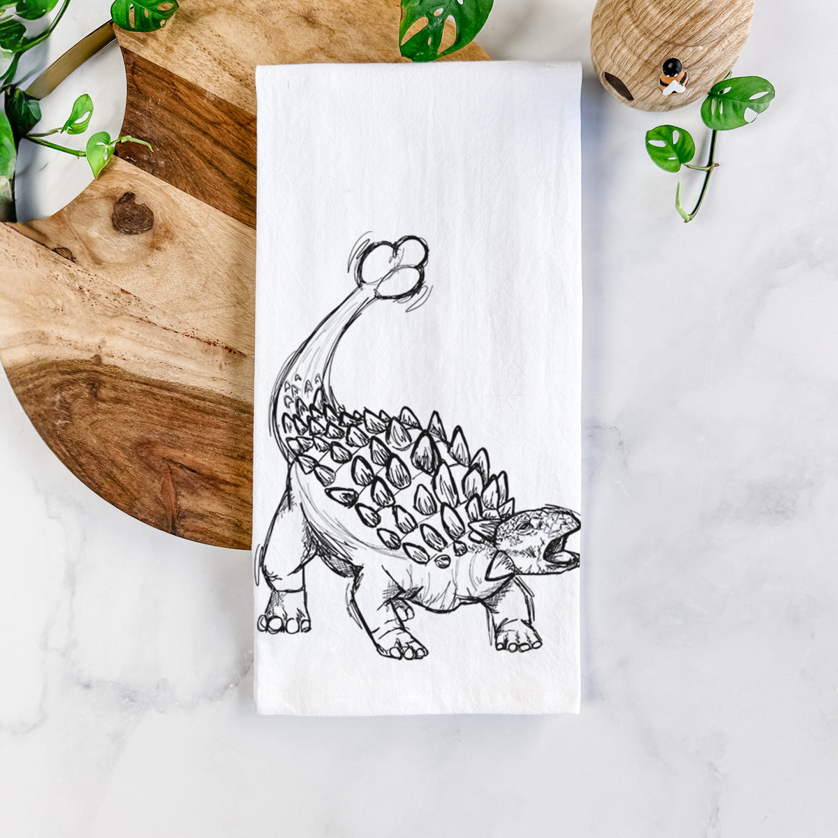 White cotton tea towel printed with a black ink illustration of an Ankylosaurus with its club tail raised, on a marble counter beside a round wooden cutting board and greenery.