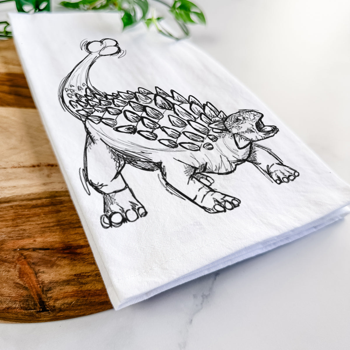 Close-up of a folded white tea towel on a wooden board, showing a black ink illustration of an Ankylosaurus with its club tail raised.