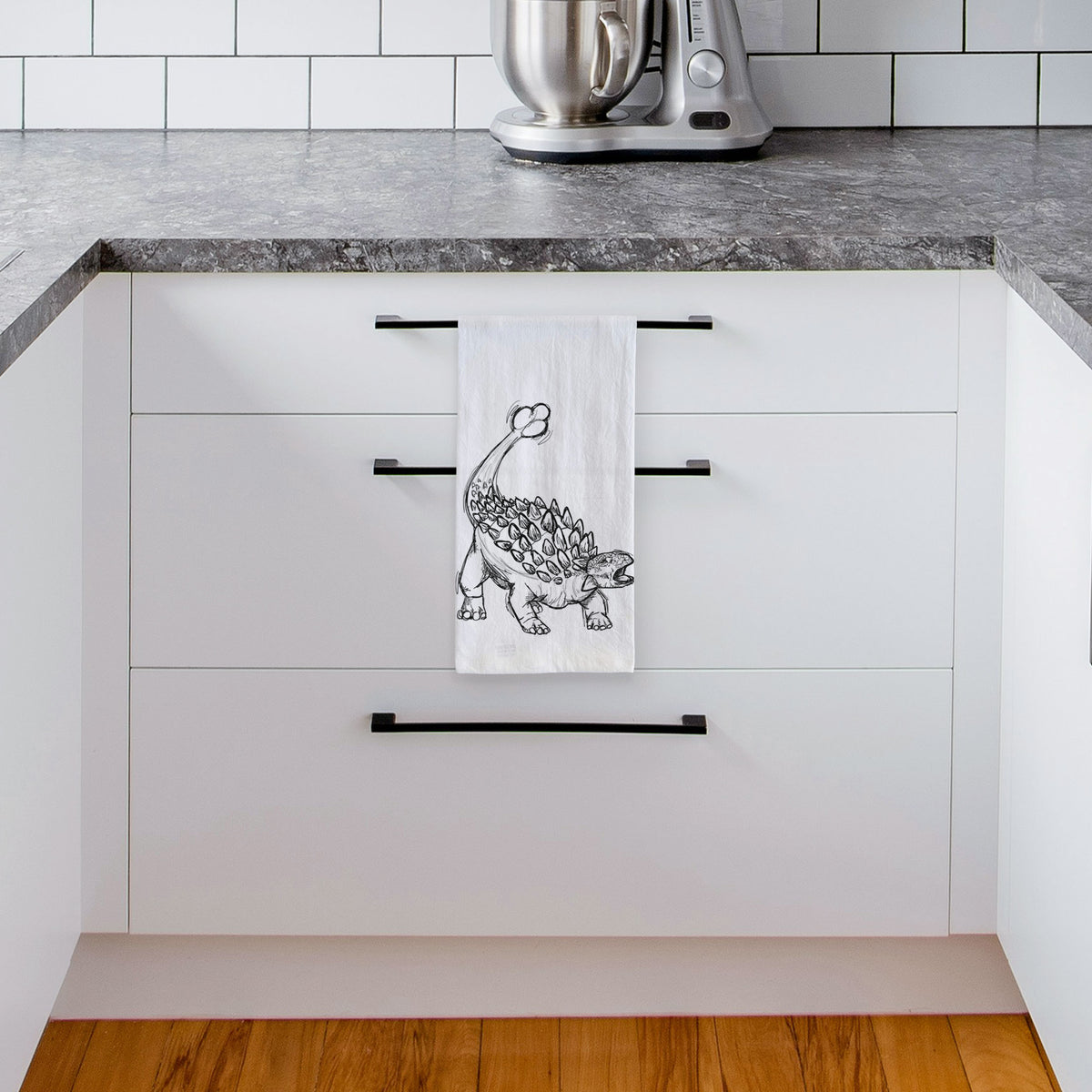 White tea towel printed with a black ink illustration of an Ankylosaurus with its club tail raised, hanging from a drawer handle in a bright white kitchen.