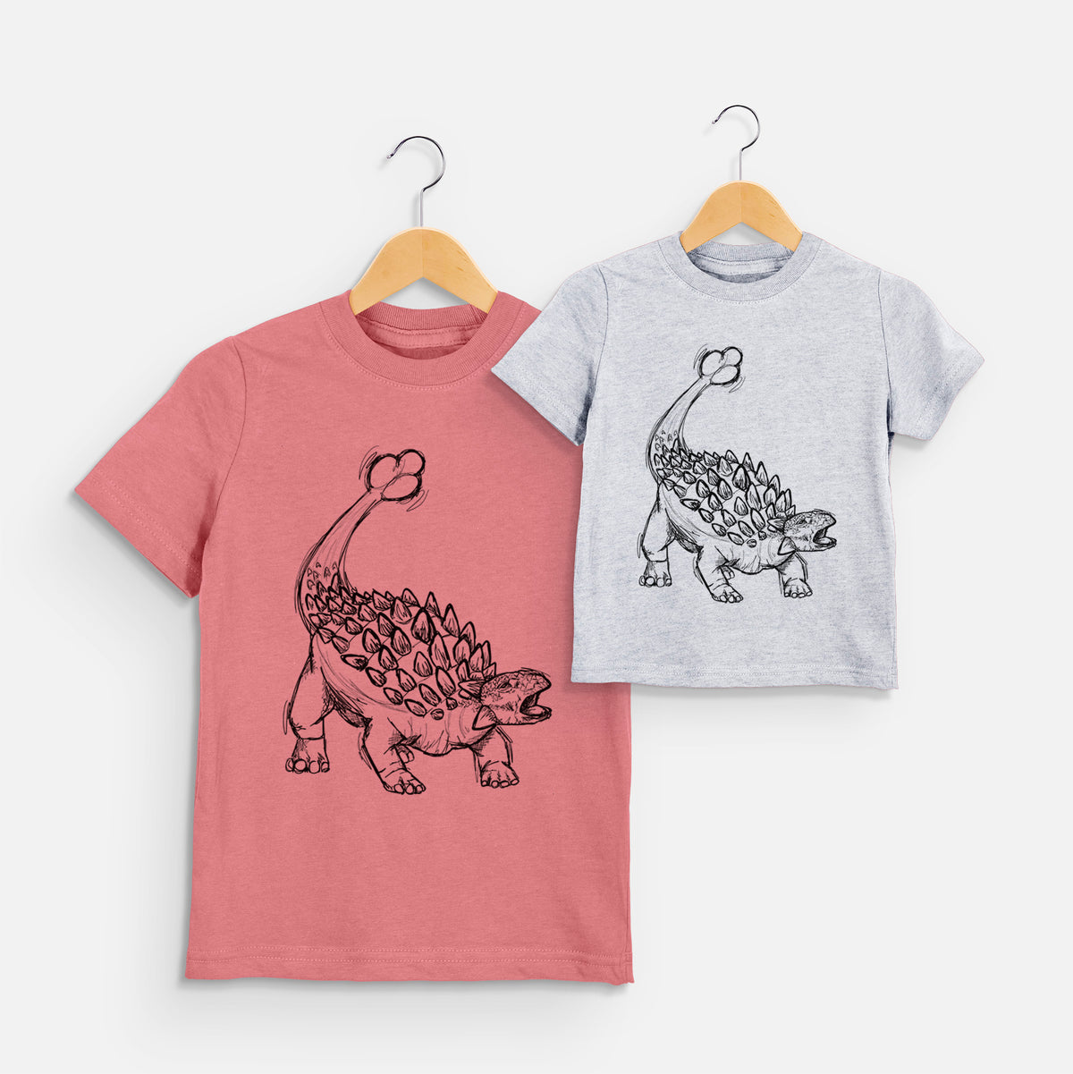 Two youth t-shirts in different colors on hangers, printed with a black ink illustration of an Ankylosaurus with its club tail raised.