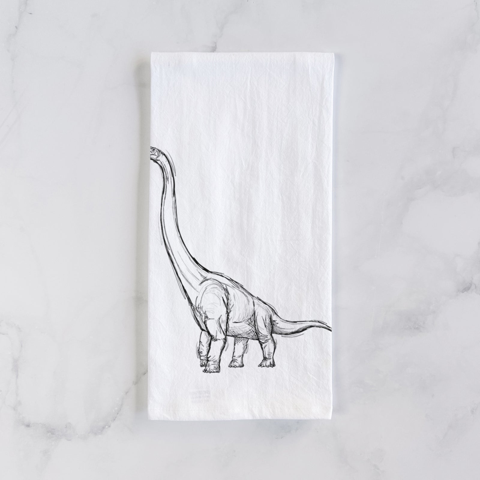 White cotton tea towel printed with a black ink illustration of a long-necked Apatosaurus walking, on a marble counter beside a round wooden cutting board and greenery.