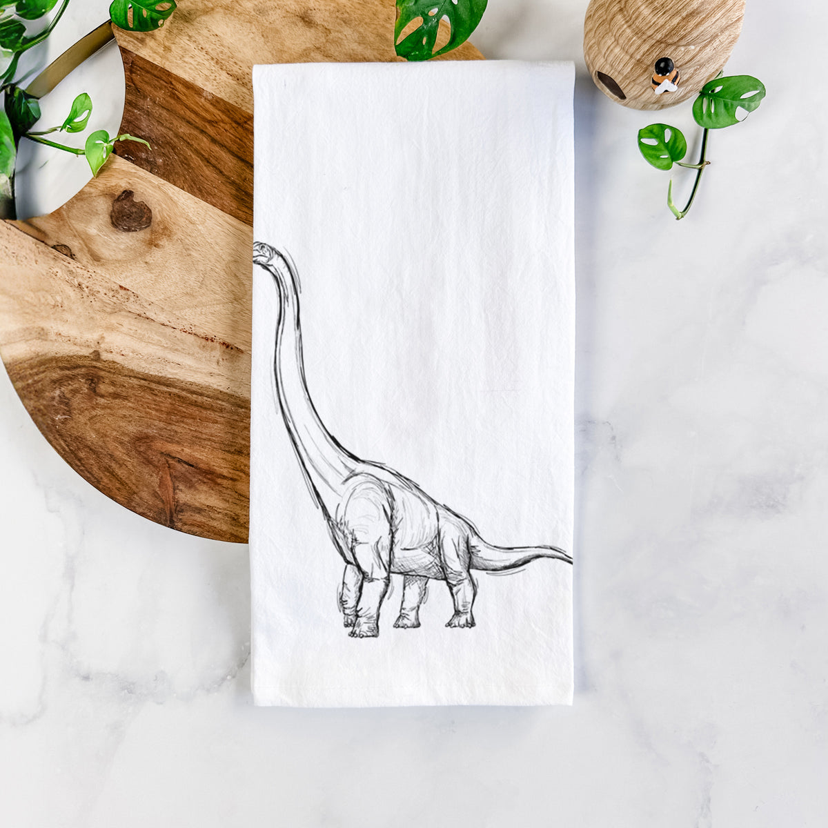 White cotton tea towel printed with a black ink illustration of a long-necked Apatosaurus walking, on a marble counter beside a round wooden cutting board and greenery.