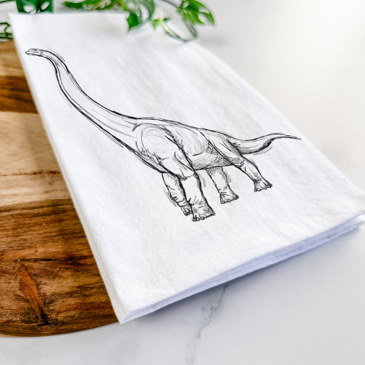 Close-up of a folded white tea towel on a wooden board, showing a black ink illustration of a long-necked Apatosaurus walking.