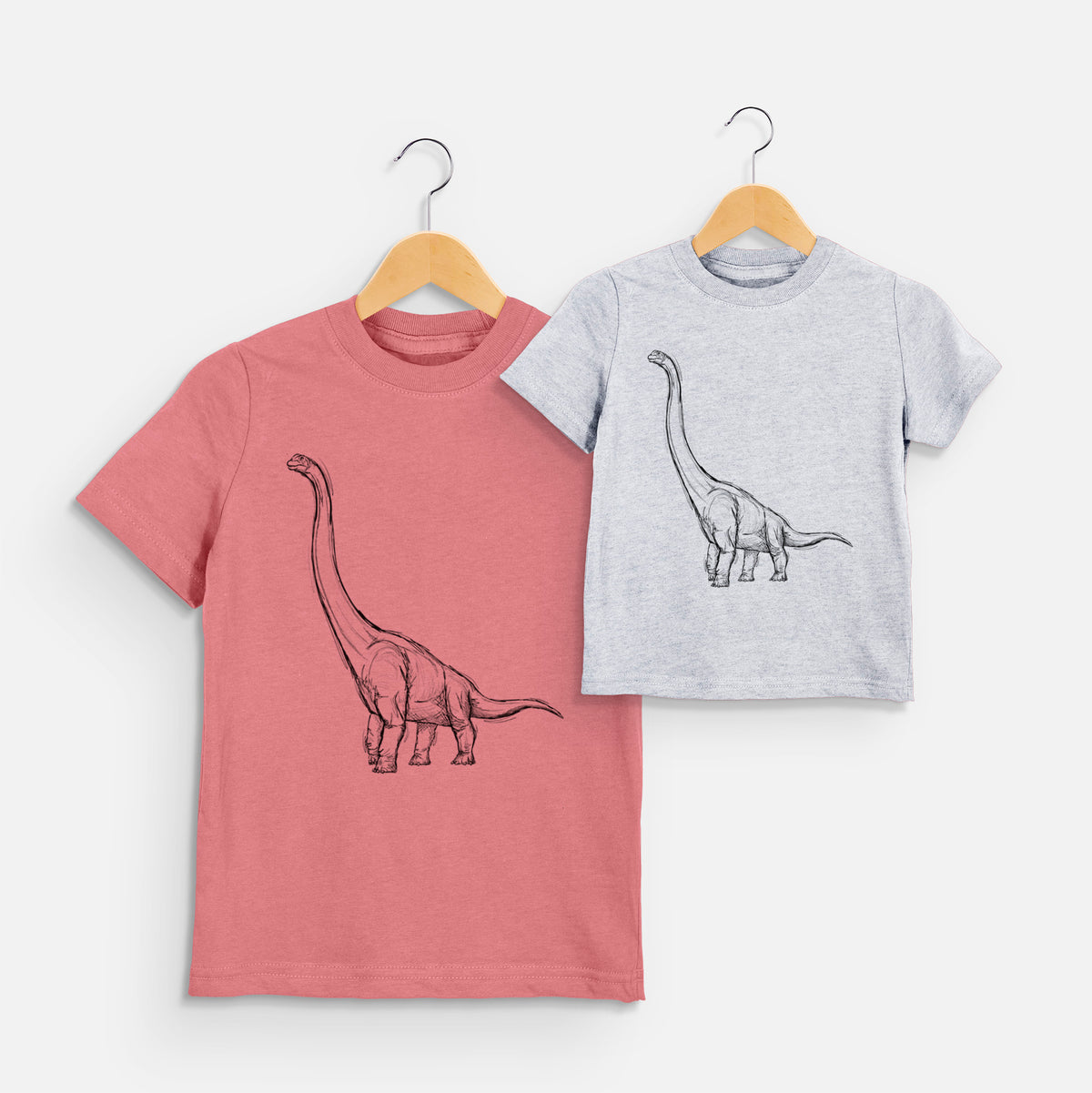 Two youth t-shirts in different colors on hangers, printed with a black ink illustration of a long-necked Apatosaurus walking.
