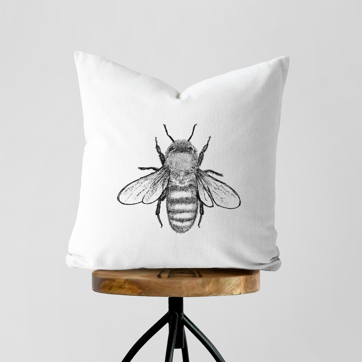 White pillow cover printed with a detailed black ink illustration of a honey bee with its wings spread, seen from above, on a wooden stool.