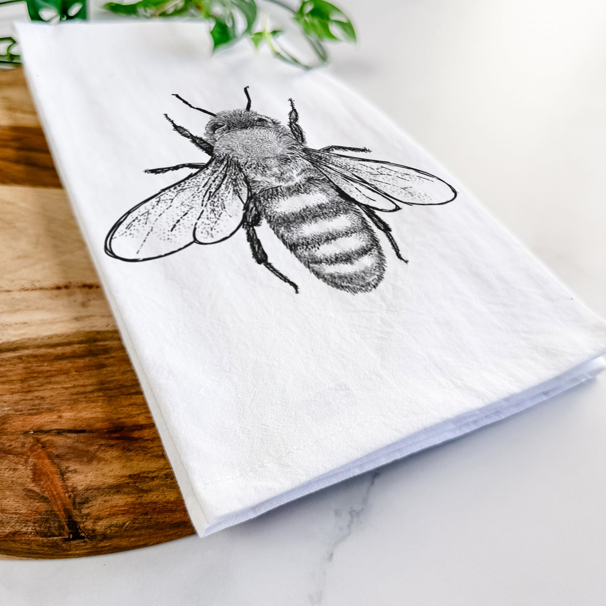 Close-up of a folded white tea towel on a wooden board, showing a detailed black ink illustration of a honey bee with its wings spread, seen from above.