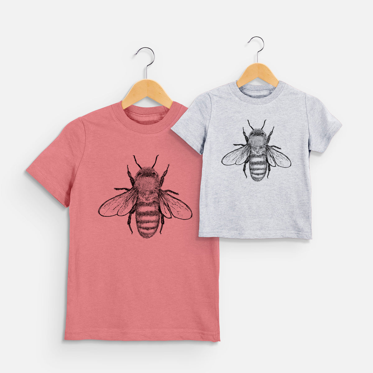 Two youth t-shirts in different colors on hangers, printed with a detailed black ink illustration of a honey bee with its wings spread, seen from above.