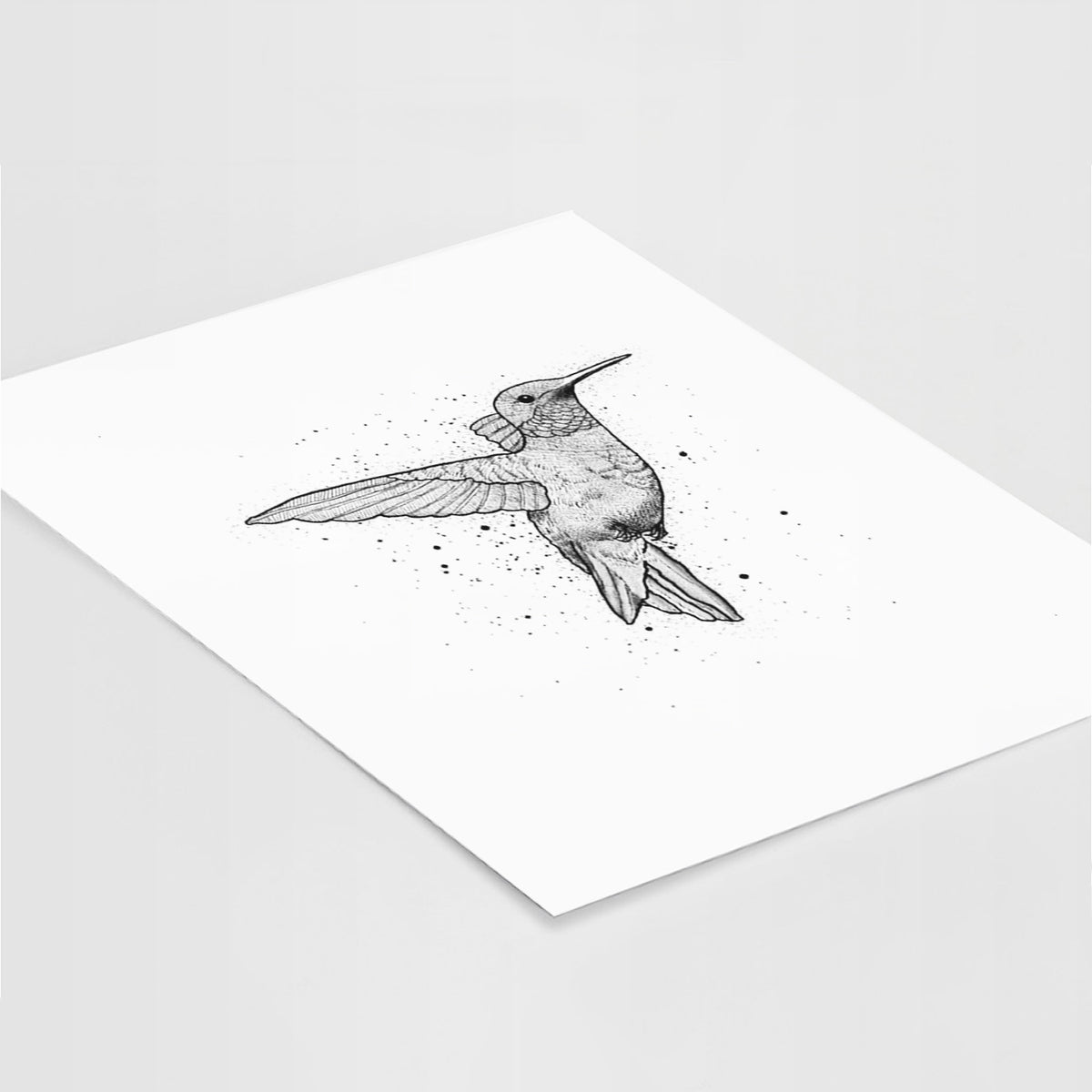 Art print of a black ink sketch of a ruby-throated hummingbird hovering with its wings raised, lying at an angle on a white surface.