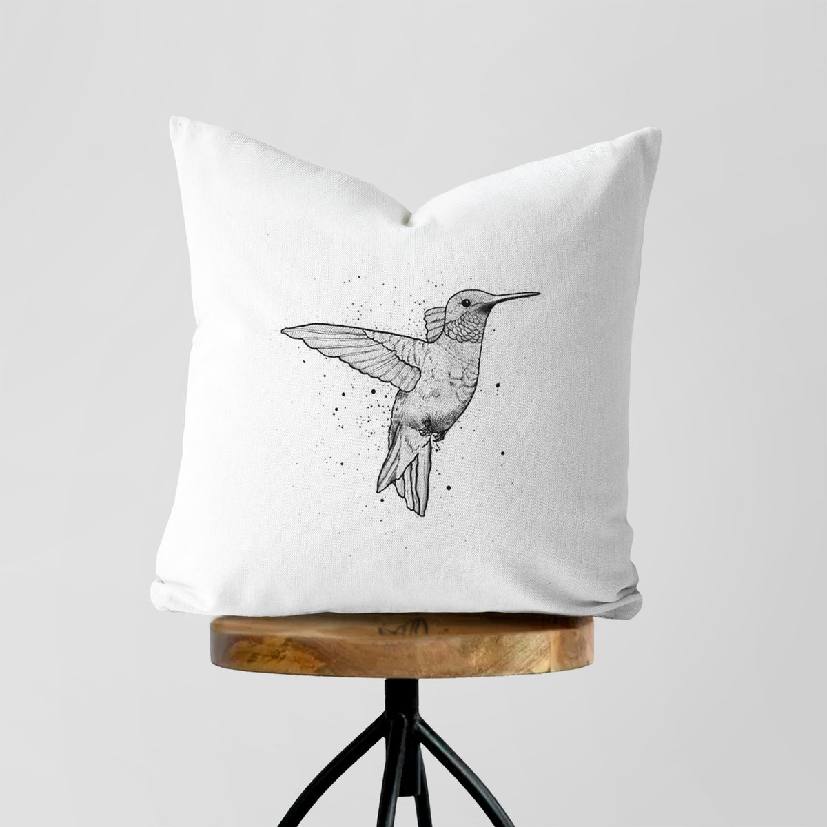 White pillow cover printed with a black ink sketch of a ruby-throated hummingbird hovering with its wings raised, on a wooden stool.