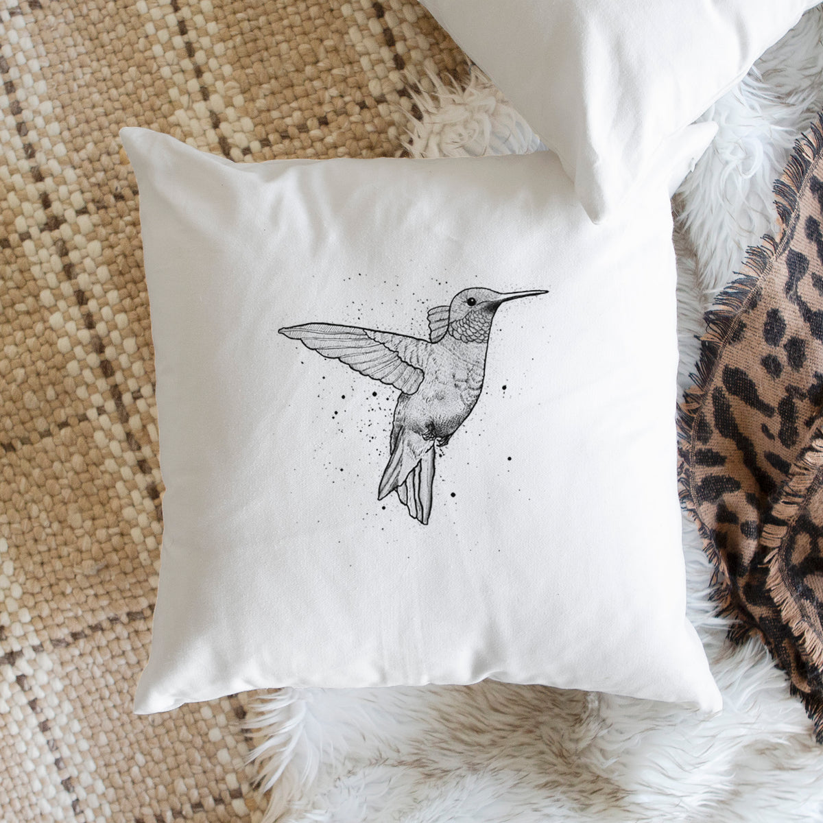 White pillow cover printed with a black ink sketch of a ruby-throated hummingbird hovering with its wings raised, on a woven chair.