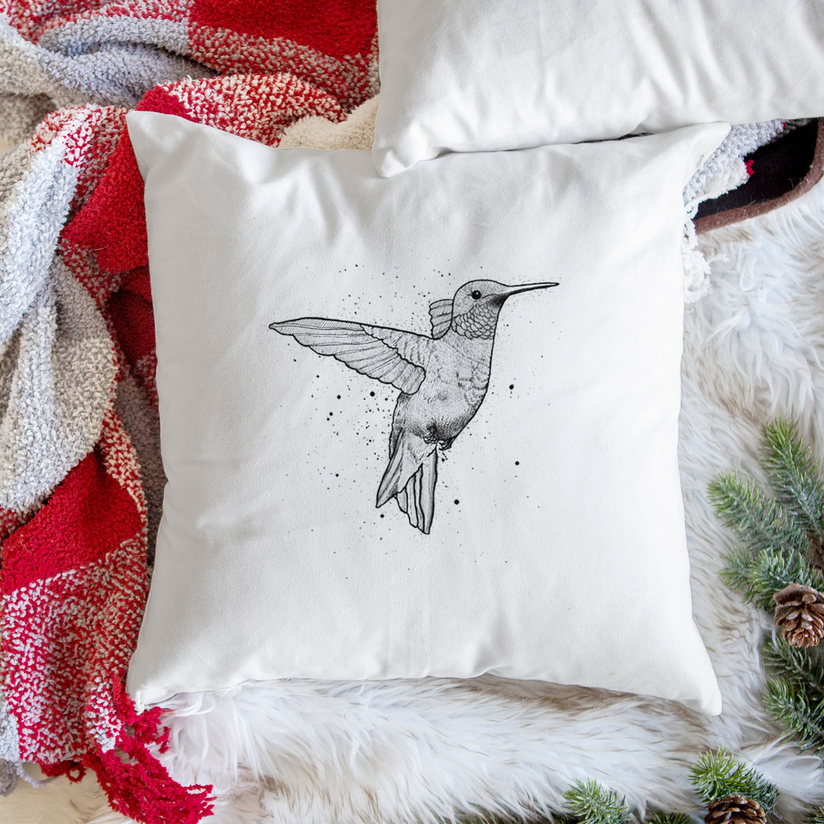 White pillow cover printed with a black ink sketch of a ruby-throated hummingbird hovering with its wings raised, on a woven chair.