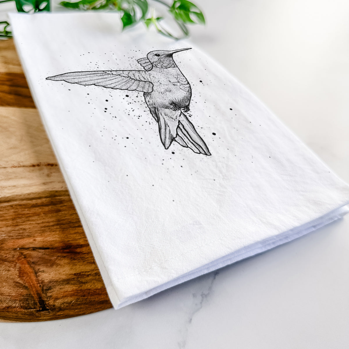 Close-up of a folded white tea towel on a wooden board, showing a black ink sketch of a ruby-throated hummingbird hovering with its wings raised.