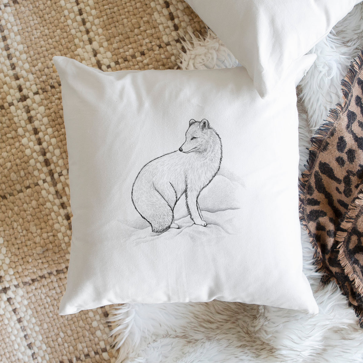White pillow cover printed with a black ink illustration of an arctic fox sitting in the snow, on a woven chair.