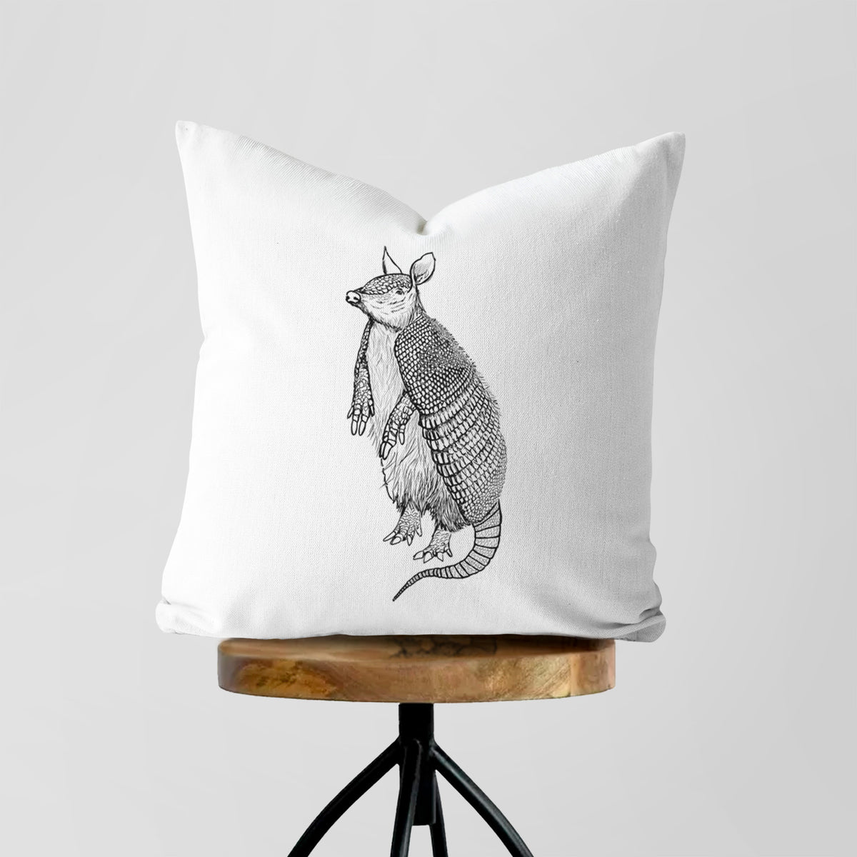 White pillow cover printed with a black ink illustration of a nine-banded armadillo standing upright, on a wooden stool.