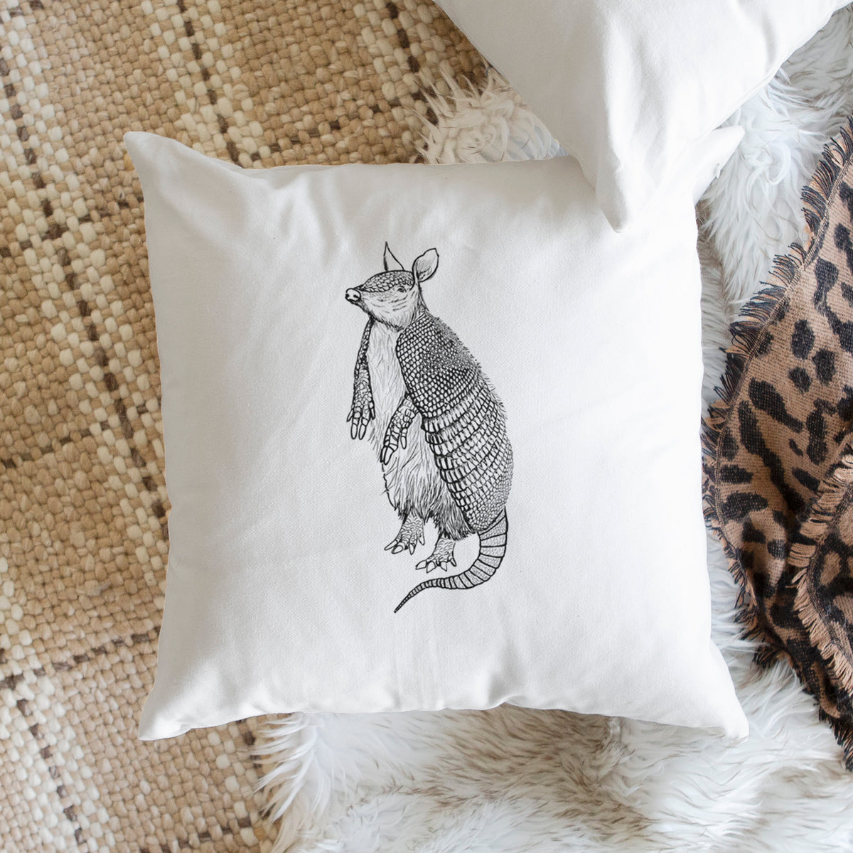 White pillow cover printed with a black ink illustration of a nine-banded armadillo standing upright, on a woven chair.