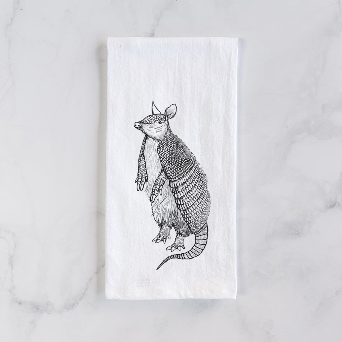 White cotton tea towel laid flat on marble, printed with a black ink illustration of a nine-banded armadillo standing upright.