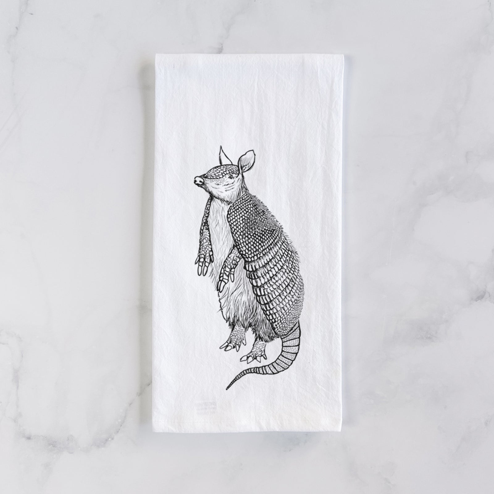 White cotton tea towel printed with a black ink illustration of a nine-banded armadillo standing upright, on a marble counter beside a round wooden cutting board and greenery.
