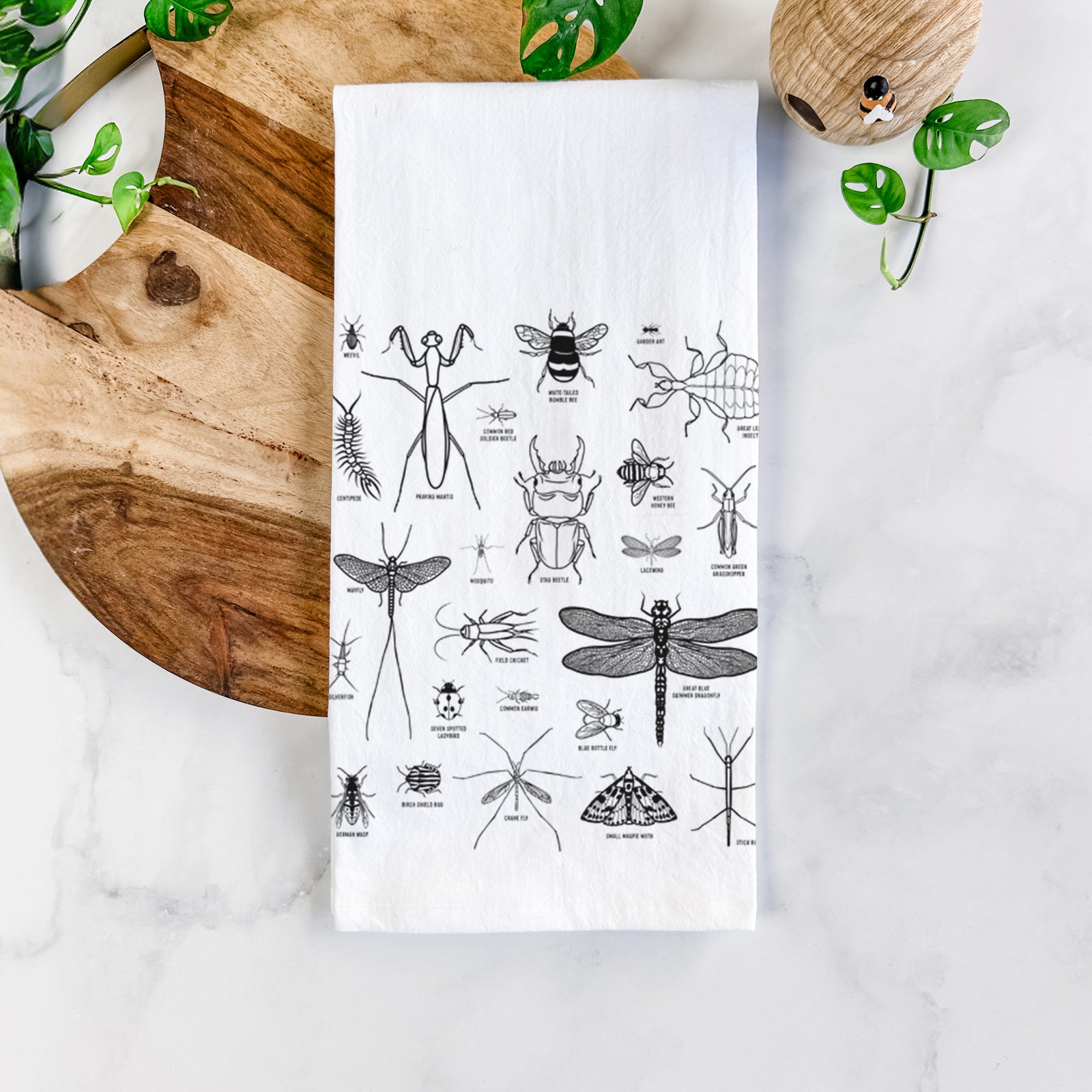 White cotton tea towel printed with a chart of black ink insect and arthropod illustrations, including dragonflies, beetles, spiders and bees, on a marble counter beside a round wooden cutting board and greenery.