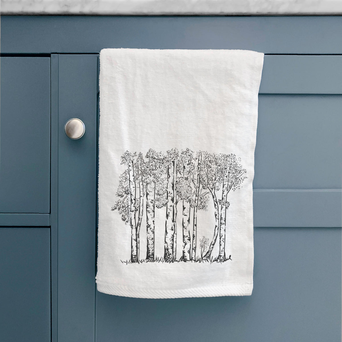 White hand towel printed with a black ink illustration of a grove of aspen trees, hanging on a cabinet door.