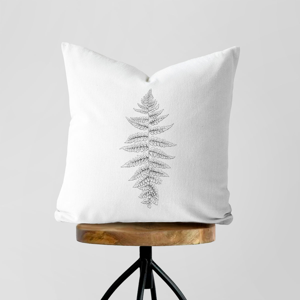 White pillow cover printed with a black ink illustration of a lady fern frond, on a wooden stool.