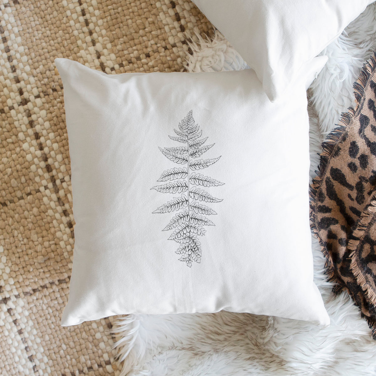 White pillow cover printed with a black ink illustration of a lady fern frond, on a woven chair.