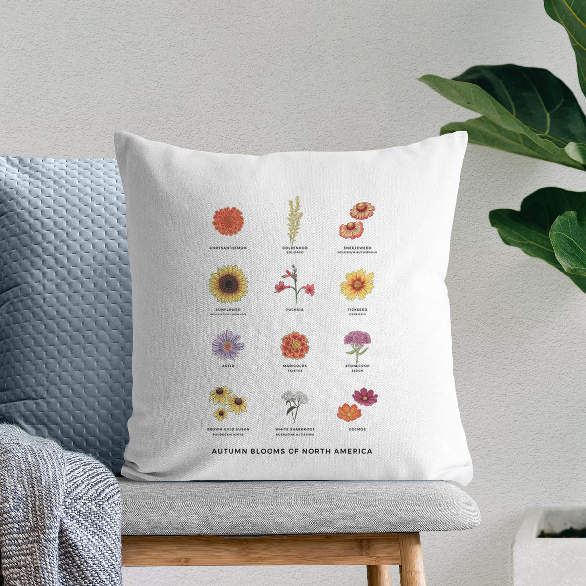 White pillow cover printed with a full-color chart of autumn flowers, each labeled with its name, on a bench beside plants.