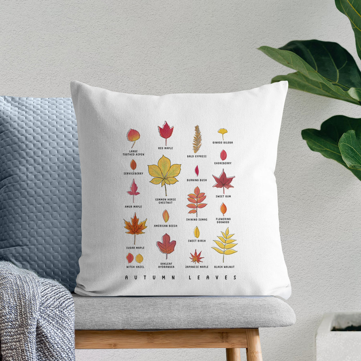 White pillow cover printed with a full-color chart of autumn leaves, each labeled with its tree species, on a bench beside plants.