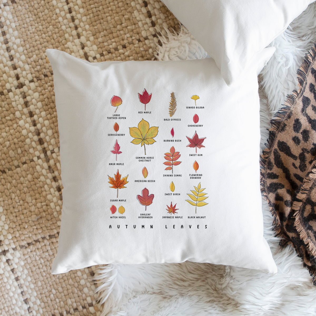 White pillow cover printed with a full-color chart of autumn leaves, each labeled with its tree species, on a woven chair.