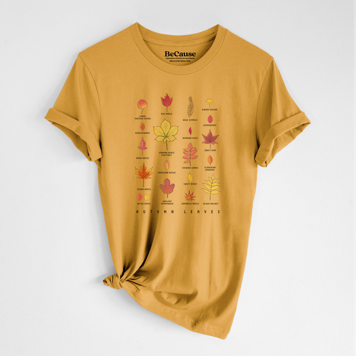 Mustard yellow lightweight cotton t-shirt laid flat with the sleeves rolled, printed with a full-color chart of autumn leaves, each labeled with its tree species.