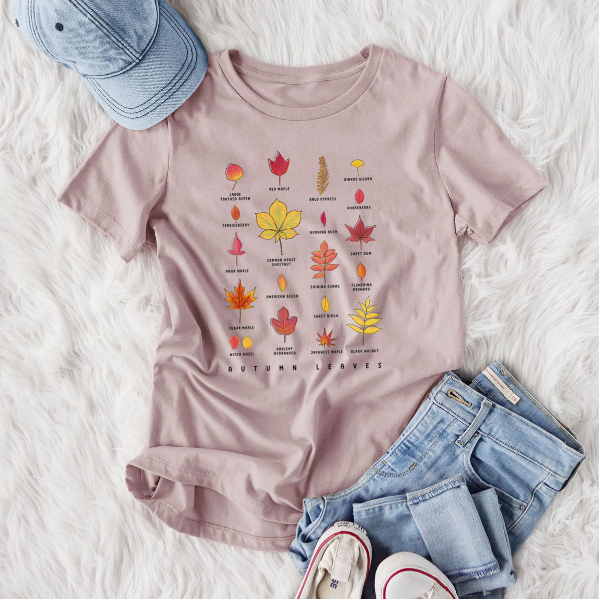Women's relaxed fit cotton t-shirt printed with a full-color chart of autumn leaves, each labeled with its tree species, styled flat on a white faux fur rug with jeans.