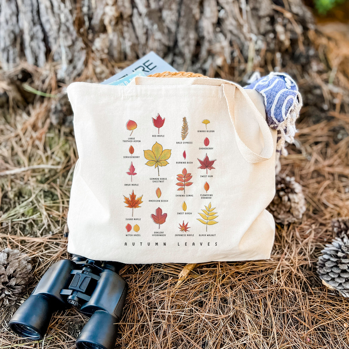 Canvas tote bag printed with a full-color chart of autumn leaves, each labeled with its tree species, resting on dry grass beside a pair of binoculars.