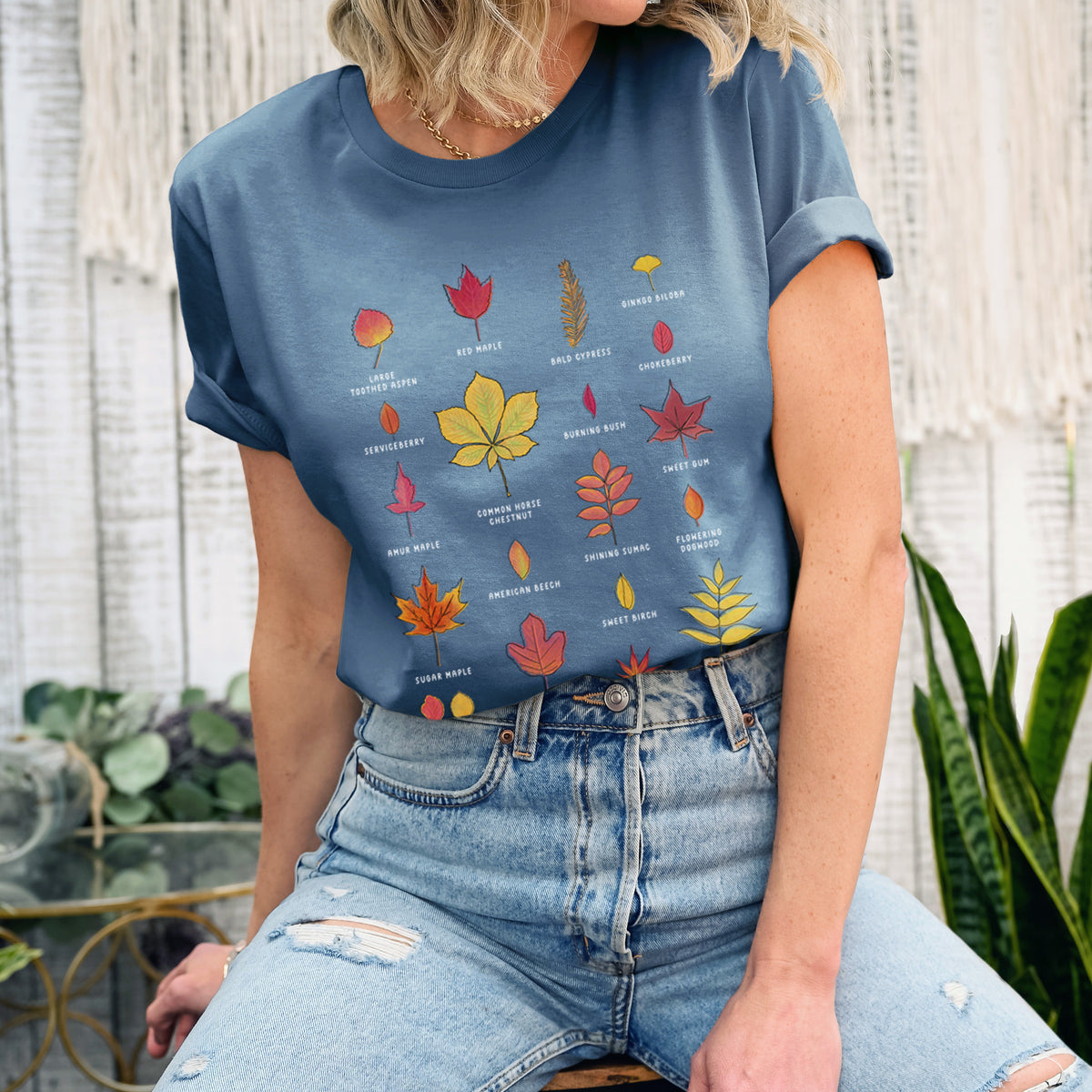 Woman wearing a steel blue lightweight cotton t-shirt printed with a full-color chart of autumn leaves, each labeled with its tree species.