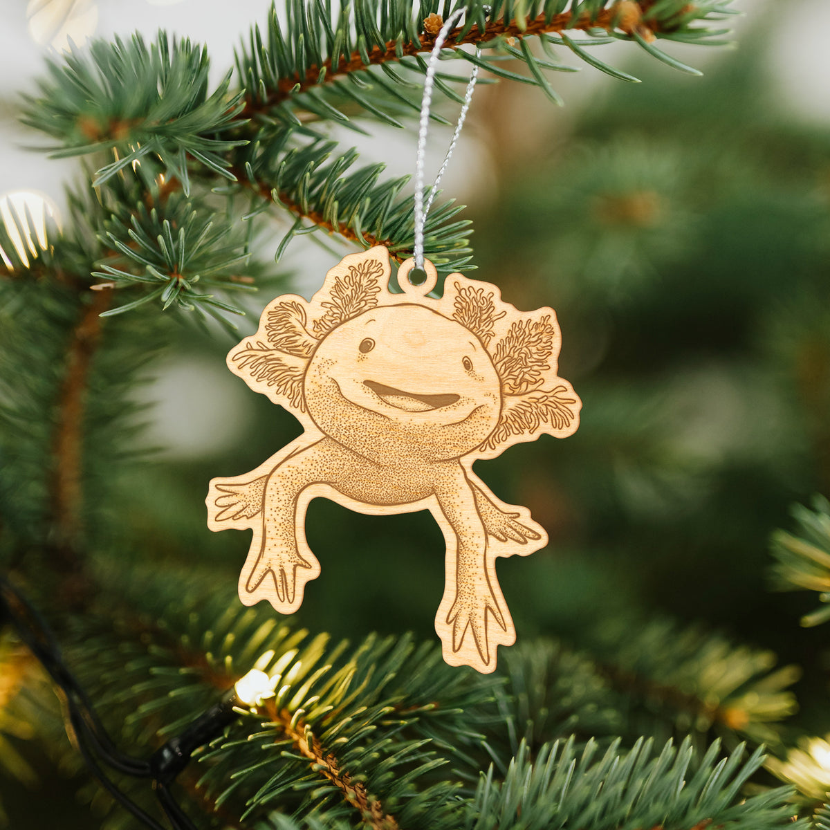 Wooden ornament featuring a engraving of a smiling axolotl with feathery gills, hanging on a Christmas tree.