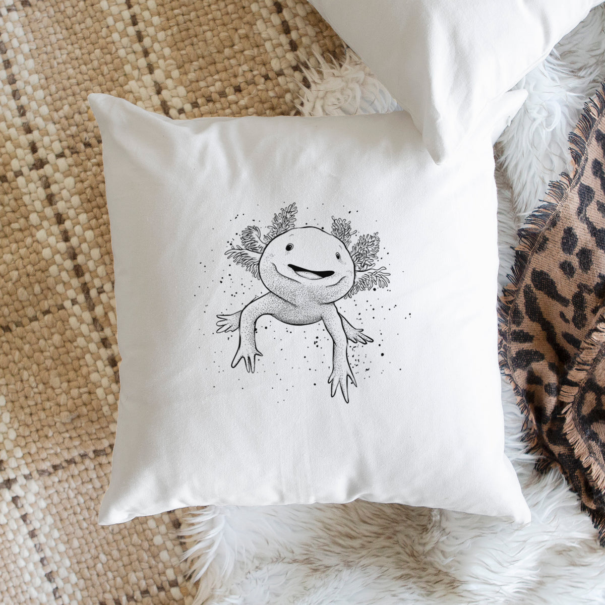 White pillow cover printed with a black ink illustration of a smiling axolotl with feathery gills, on a woven chair.