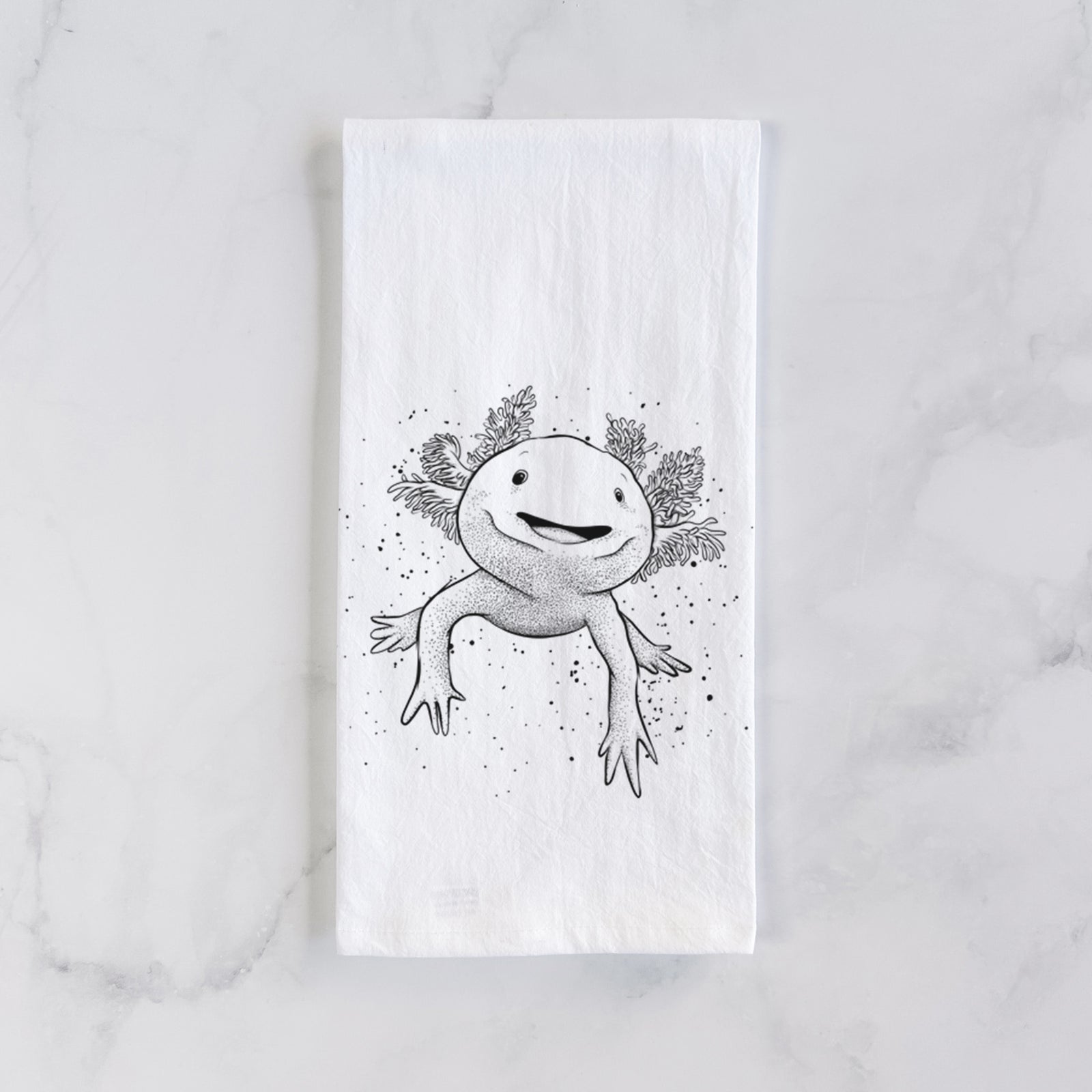 White cotton tea towel printed with a black ink illustration of a smiling axolotl with feathery gills, on a marble counter beside a round wooden cutting board and greenery.