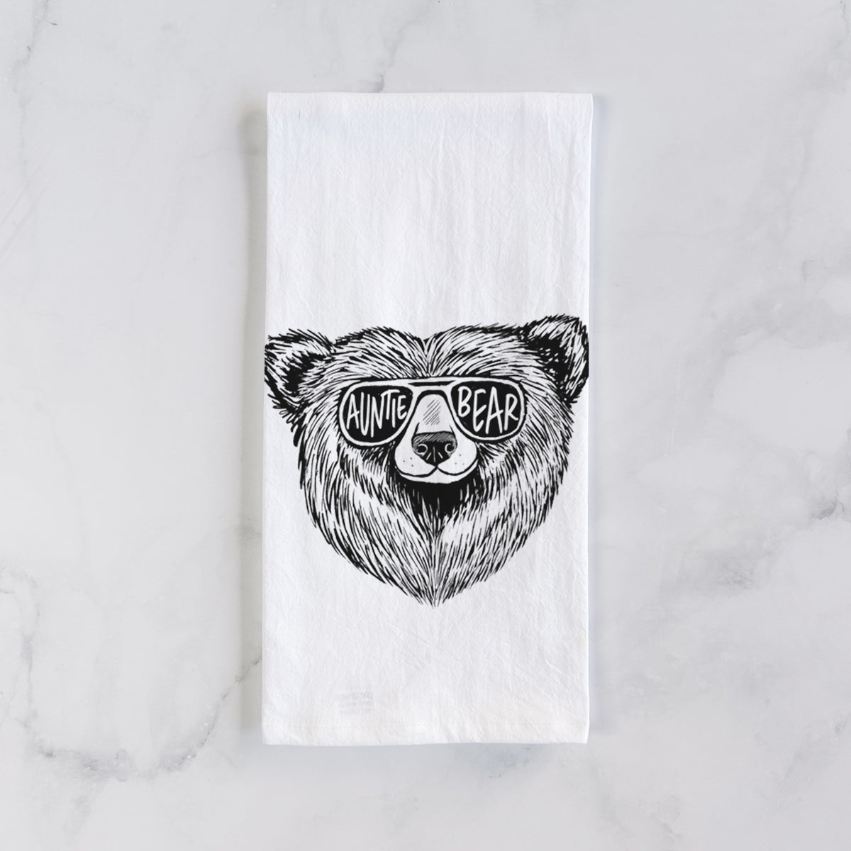 White cotton tea towel laid flat on marble, printed with a black ink bear's face wearing sunglasses that read Auntie Bear.