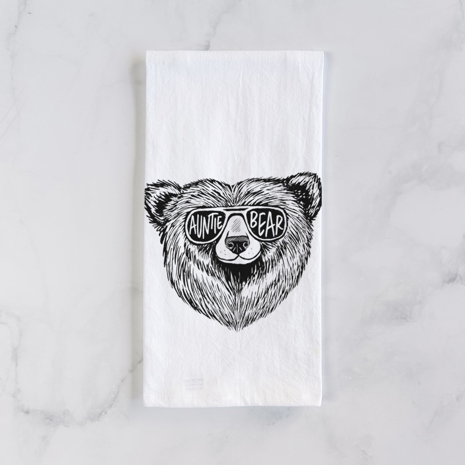 White cotton tea towel printed with a black ink bear's face wearing sunglasses that read Auntie Bear, on a marble counter beside a round wooden cutting board and greenery.