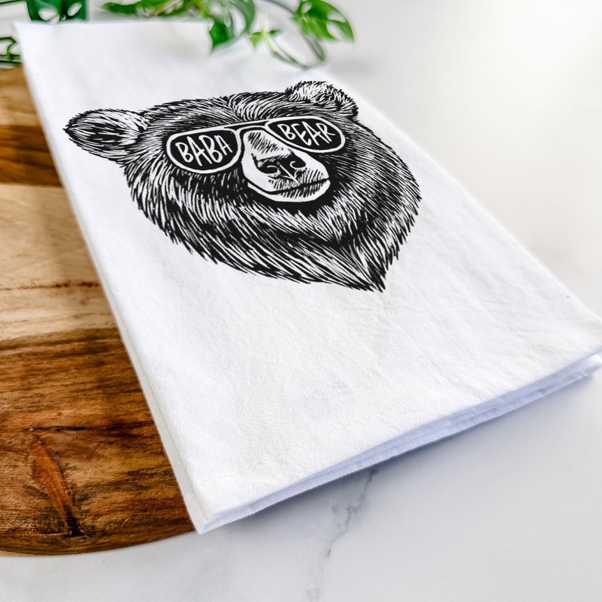 Close-up of a folded white tea towel on a wooden board, showing a black ink bear's face wearing sunglasses that read Baba Bear.