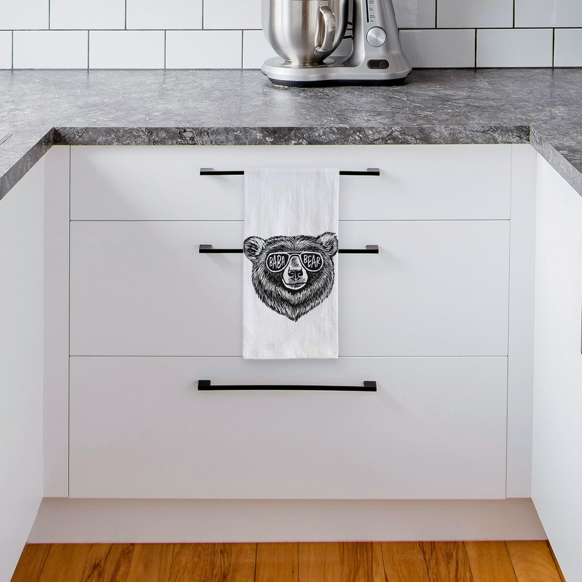 White tea towel printed with a black ink bear's face wearing sunglasses that read Baba Bear, hanging from a drawer handle in a bright white kitchen.