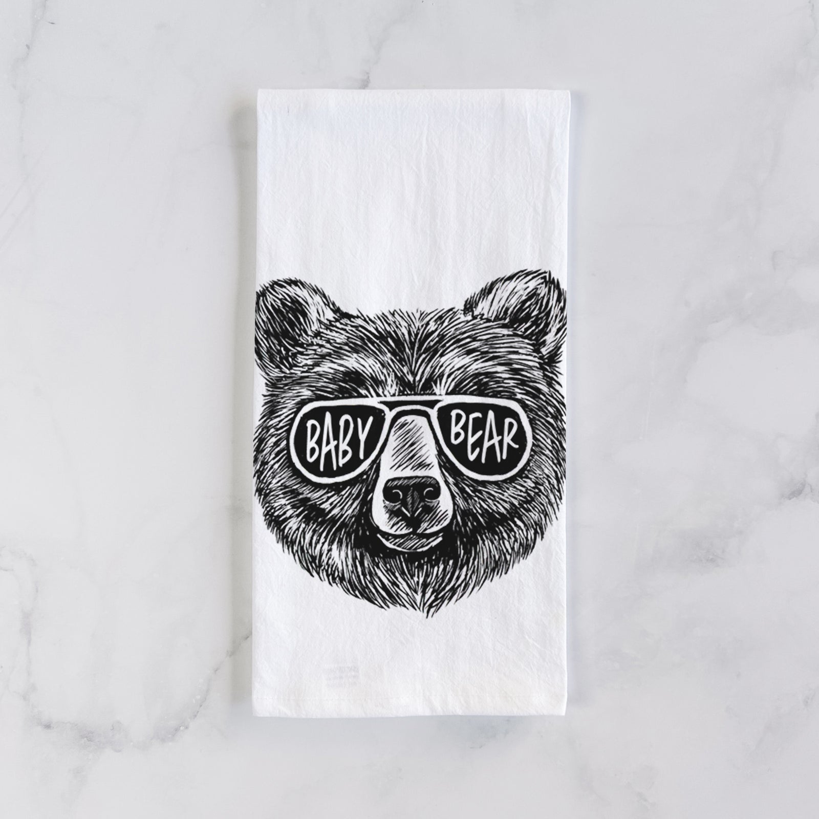 White cotton tea towel printed with a black ink bear's face wearing sunglasses that read Baby Bear, on a marble counter beside a round wooden cutting board and greenery.