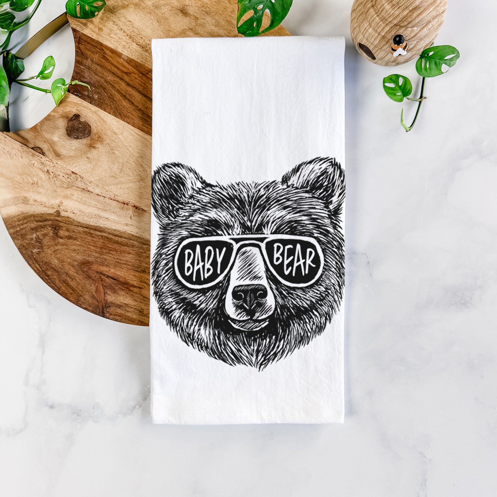 White cotton tea towel printed with a black ink bear's face wearing sunglasses that read Baby Bear, on a marble counter beside a round wooden cutting board and greenery.